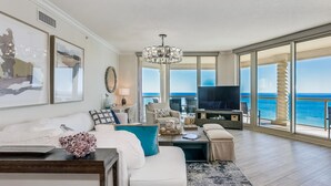 A spacious living room with a large window offering a view of the ocean, a chandelier, and a television on a wooden stand.