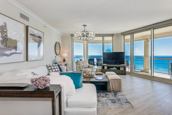 A spacious living room with a large window offering a view of the ocean, a chandelier, and a television on a wooden stand.