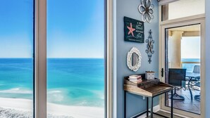 A room with a large window overlooking a beach, a wooden table with books and a decorative mirror, and wall art including a starfish and a flower.