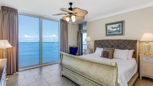 Une chambre d’hôtel avec un grand lit, un ventilateur de plafond, une vue sur l’océan et un tableau encadré au mur.