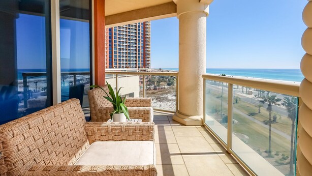 Un balcón con mobiliario de mimbre, una planta en maceta y vistas a la playa y un edificio alto.