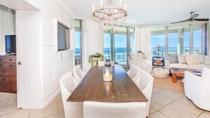 Amplia sala de estar con una gran mesa de comedor de madera, sofás blancos y una lámpara de araña. Un televisor está montado en la pared y hay vista al océano a través de amplias ventanas.