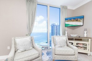 A room with two patterned armchairs, a small round table, a television mounted on the wall, and a view of the ocean and buildings through a large window.