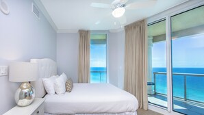 A bedroom with a bed, a nightstand, a lamp, and a view of the ocean through large windows.