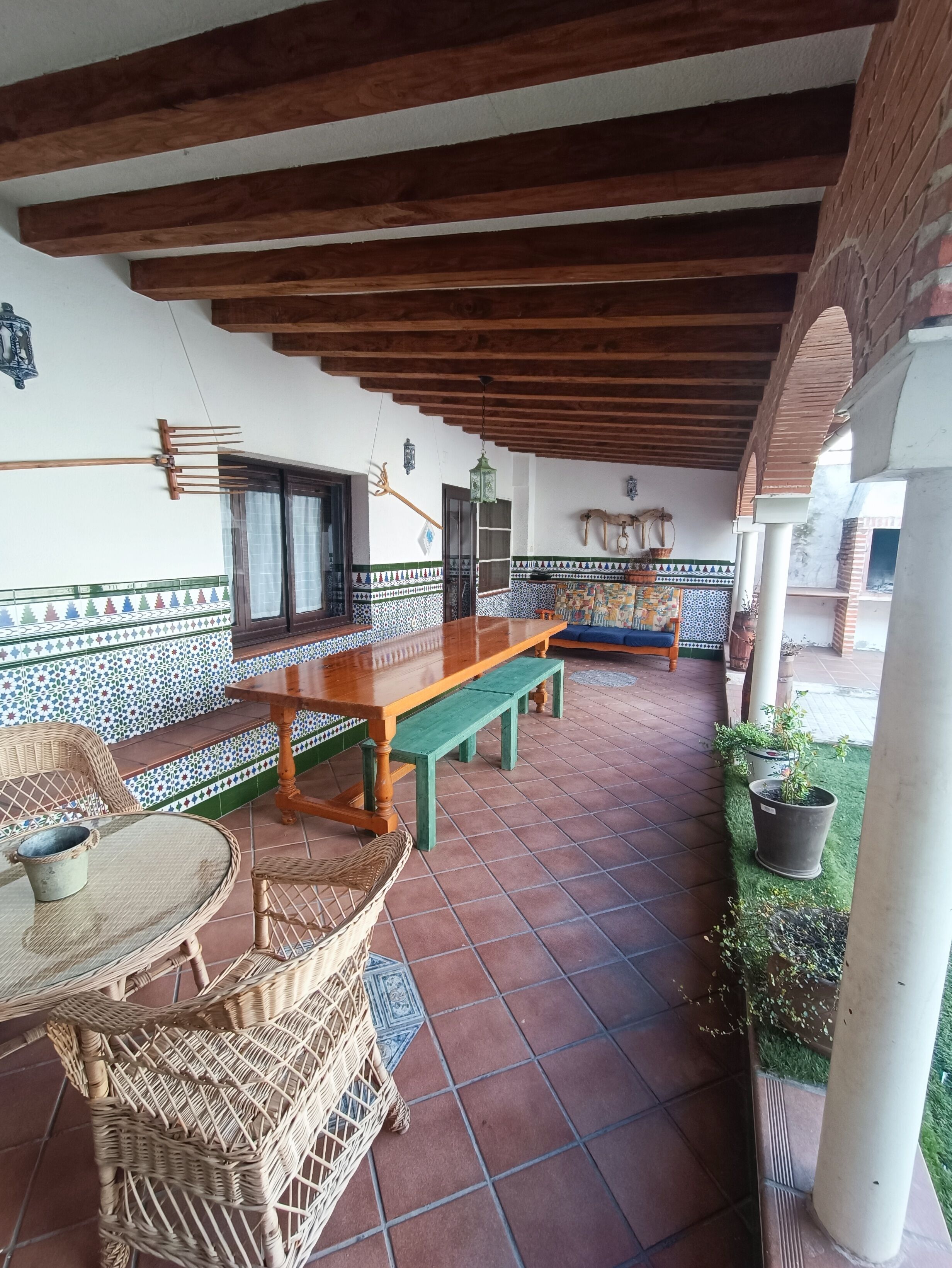 A covered patio area with a wooden table and chairs, a bench, and a tiled floor.