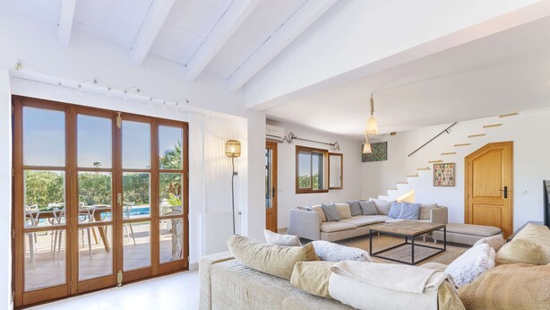 A spacious living area with a large sofa, a coffee table, and a dining area with chairs and a table. There is a staircase leading to another level, and a wooden door is visible. - Villa Miravall Selva by Balearic Villas (Selva)