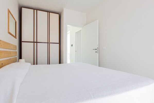 A bedroom with a large bed, a wardrobe with vertical lines, a mirror, and a door leading to another room.