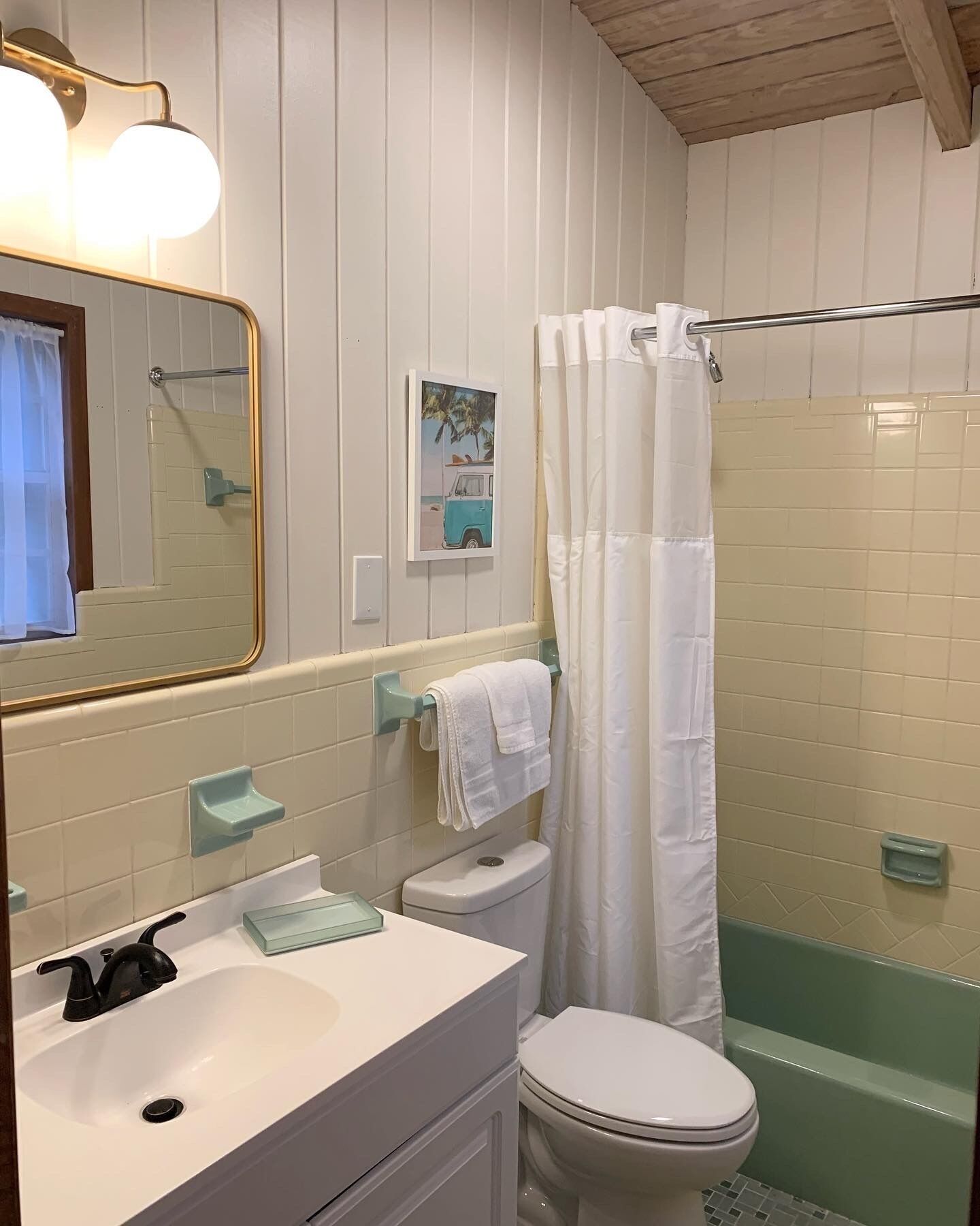Een badkamer met een witte wastafel, toilet en douche. Er is een spiegel met gouden lijst, een handdoekhouder en een foto aan de muur.