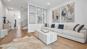 A modern living room with a light-colored sofa, a coffee table, and a large rug. There are framed artworks on the wall and a staircase leading to another room.