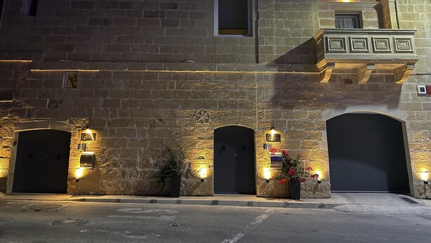 A stone building with multiple arched doorways, a small balcony with a railing, and exterior lighting. - Heritage Living Apartment With Spectacular Views (Zebbug)