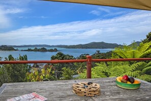Une terrasse en bois donnant sur une baie, un saladier en bois rempli de fruits et un magazine posé sur la table.
