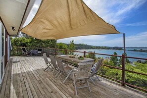 Une terrasse en bois avec une table et des chaises, un grand auvent et une vue panoramique sur un lac et des collines.