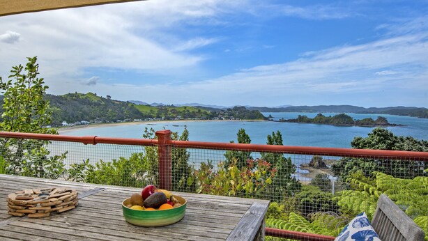 Een houten terras met een tafel, een fruitschaal en een prachtig uitzicht op een baai met heuvels en een heldere lucht.