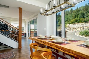 Un coin repas avec une table en bois dressée pour quatre personnes, de grandes fenêtres donnant sur un paysage boisé et un escalier avec une rampe en bois.
