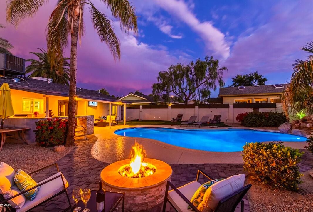 Piscina con un pozo de fuego, sillas de descanso y un patio con vistas a una casa y palmeras.