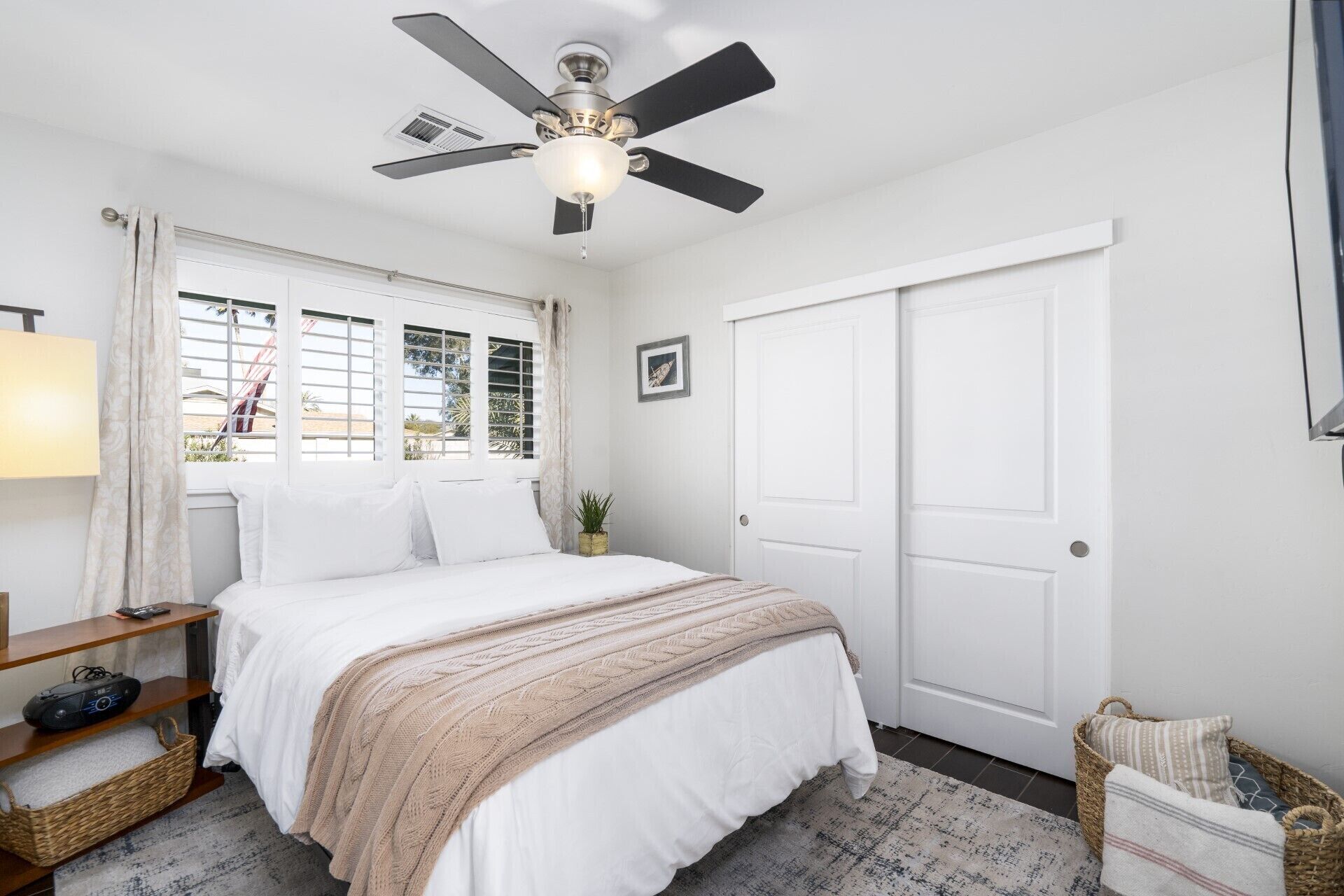 Une chambre à coucher avec un lit, un ventilateur de plafond, une fenêtre avec des rideaux, une table de chevet avec une lampe, une porte et une petite plante.