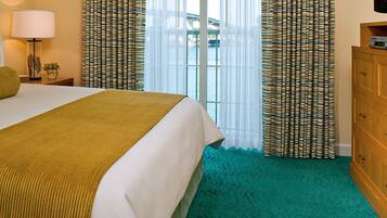 A hotel room with a bed, a wooden dresser, a lamp, and curtains.