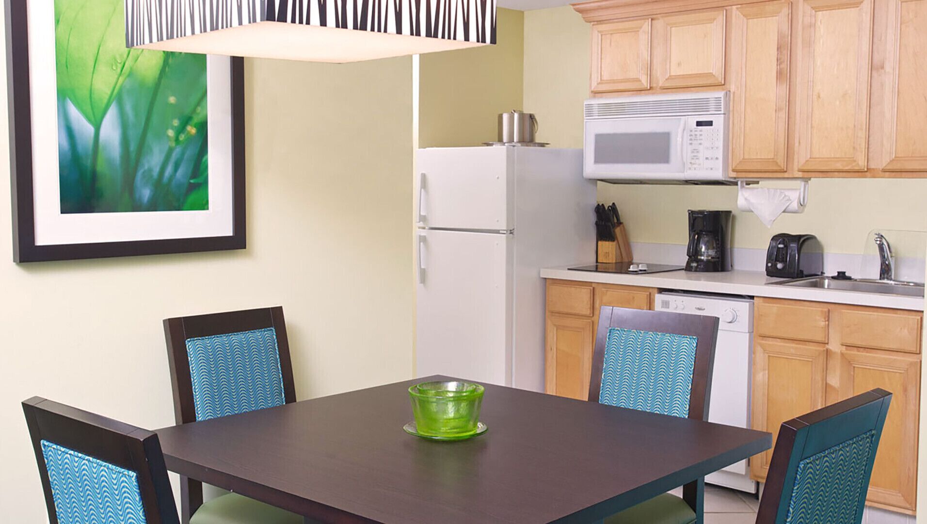 Cocina compacta con refrigerador, microondas y cafetera. Hay una mesa de comedor con sillas y un cuadro en la pared.