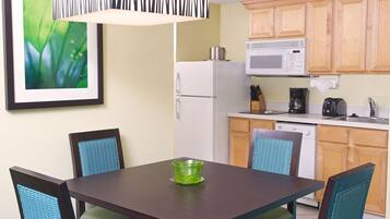 Cocina compacta con refrigerador, microondas y cafetera. Hay una mesa de comedor con sillas y un cuadro en la pared.