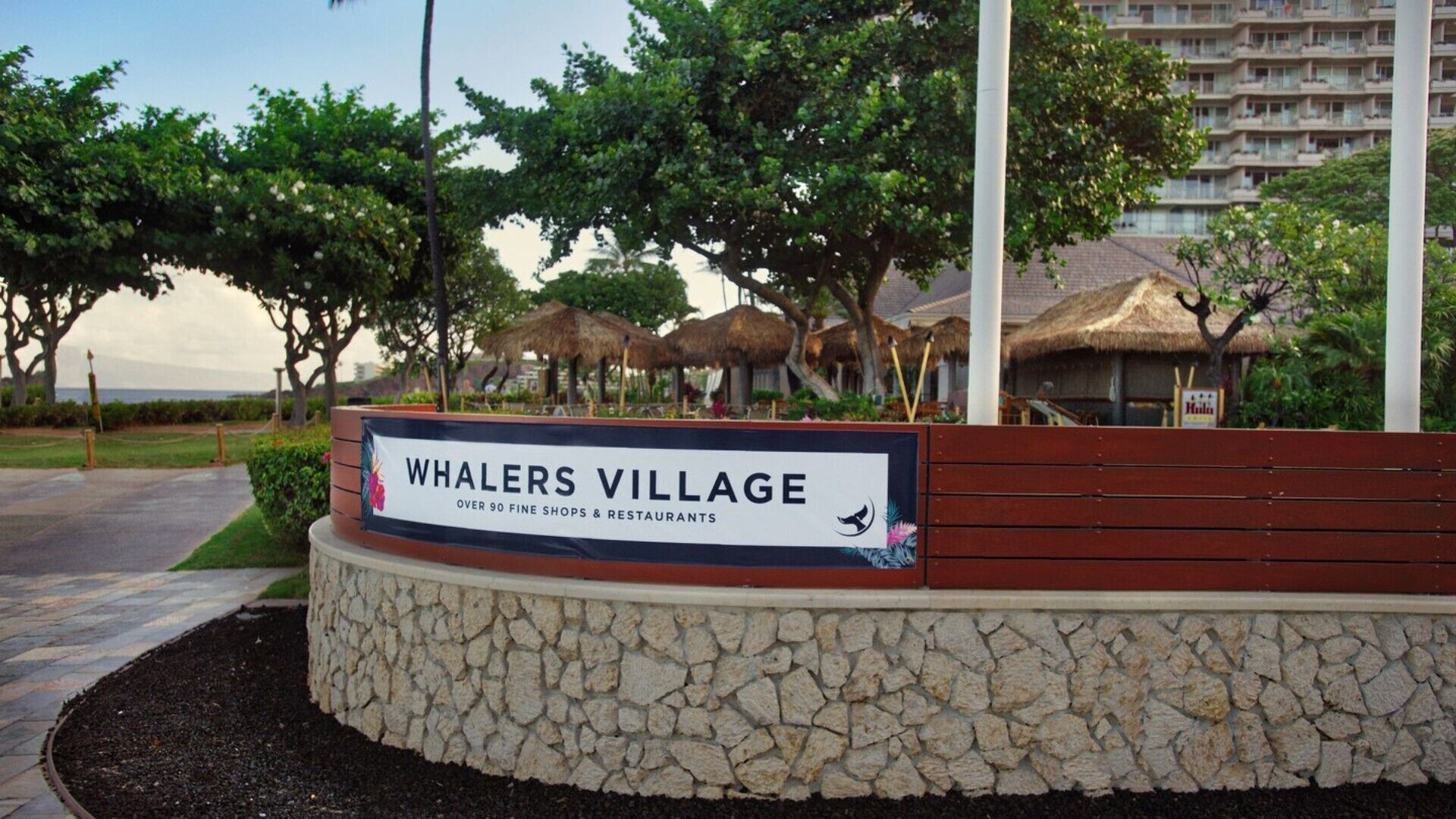 A sign for Whalers Village with a stone base and a wooden top, indicating over 90 fine shops and restaurants.