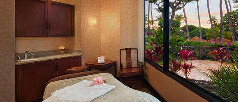 Habitación de hotel con una cama, baño con lavamanos, una silla y vista a un jardín con palmeras y flores.