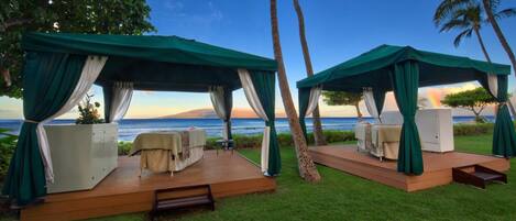 Two outdoor cabanas with beds and seating, overlooking a beach and ocean.