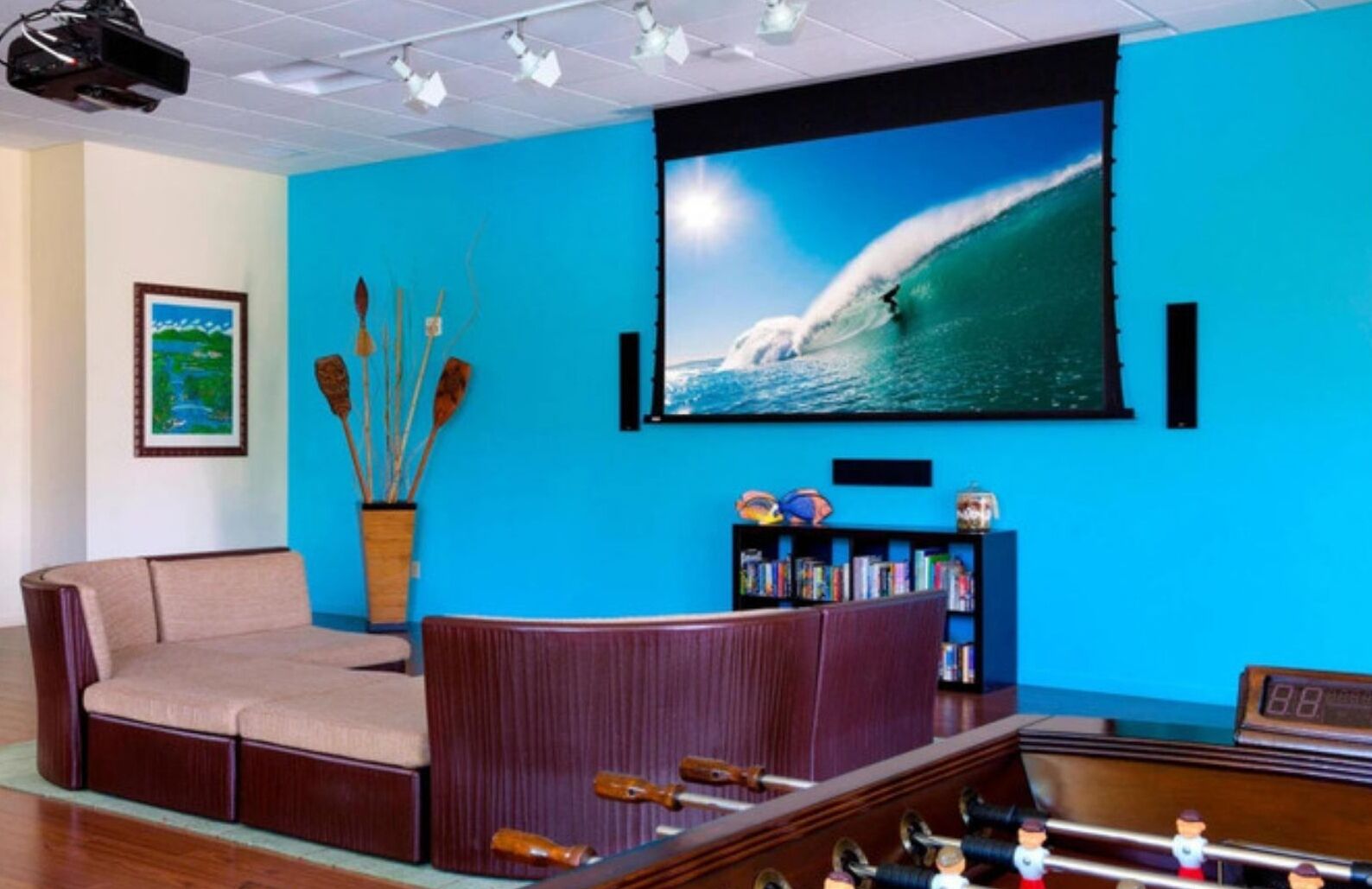 Una habitación con una pared azul, una pantalla grande de televisión mostrando una ola, un sofá, una estantería y una mesa de fútbol.