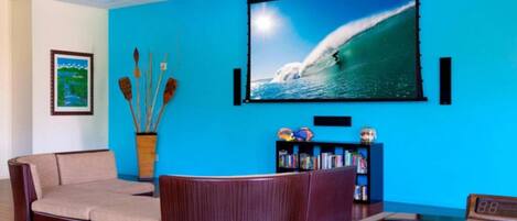 Una habitación con una pared azul, una pantalla grande de televisión mostrando una ola, un sofá, una estantería y una mesa de fútbol.