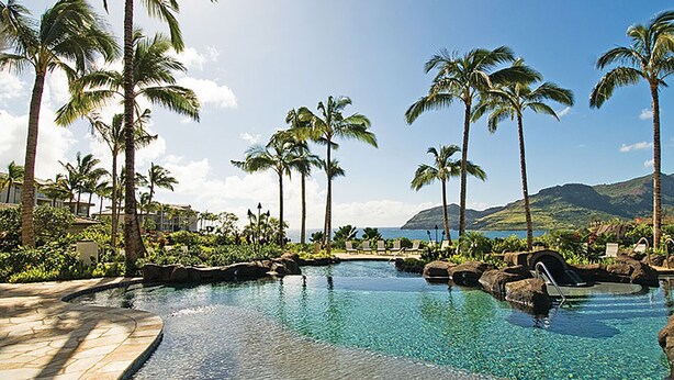 A resort pool area with clear water, surrounded by palm trees and a view of the ocean and mountains.