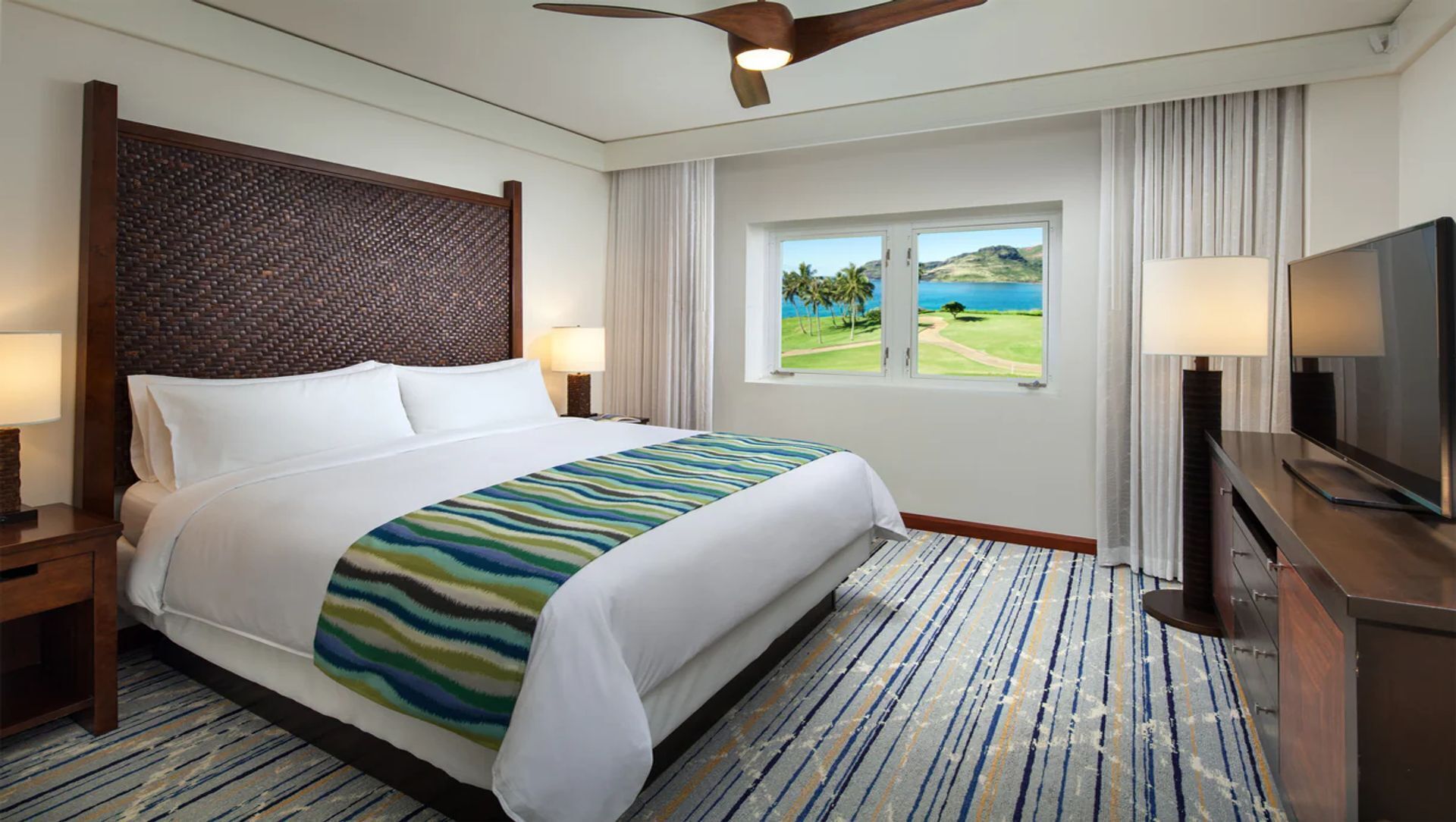 Habitación de hotel con una cama grande, ventilador de techo, televisión y ventana con vistas a un campo de golf.