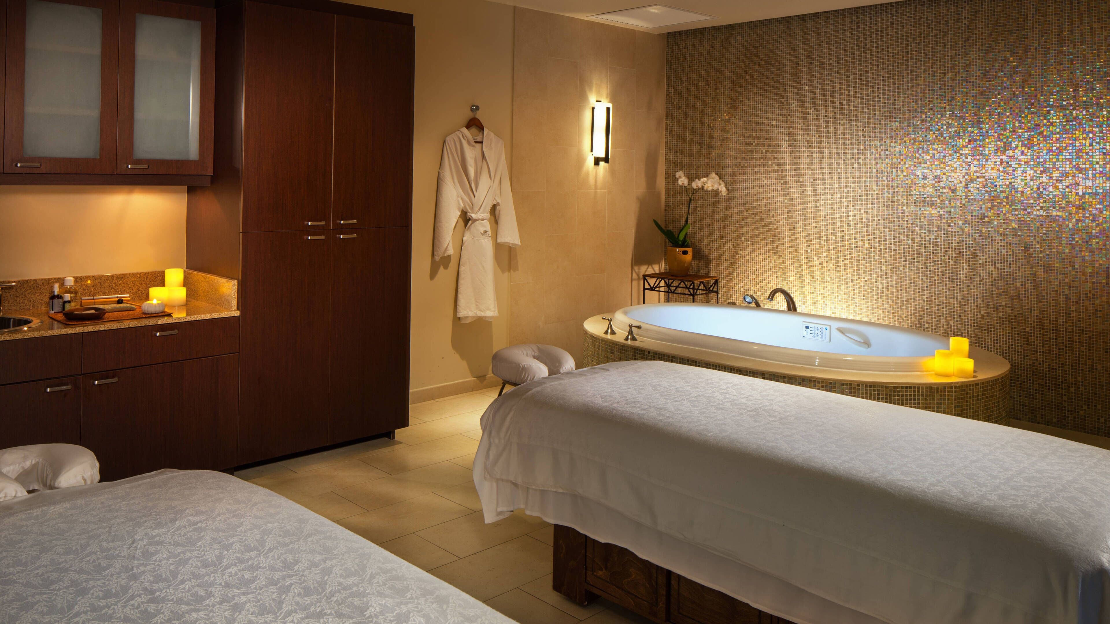 A spa room with a large bathtub, two beds, a wooden cabinet, and a wall-mounted light fixture.