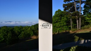 A sign with the text 'THIS IS OUR HAPPY PLACE' and a view of a road with trees and a distant mountain range. - Unwine'd With a View (Hayesville)