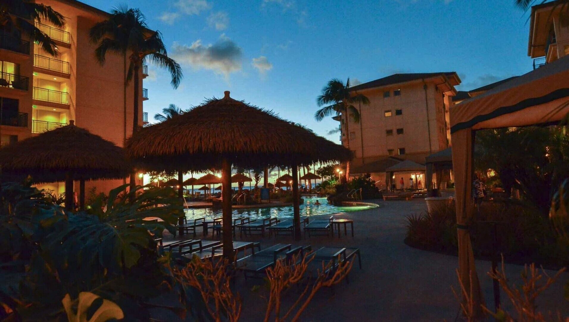 Zona de piscina en un resort, con sombrillas de paja, sillas de descanso y una piscina.