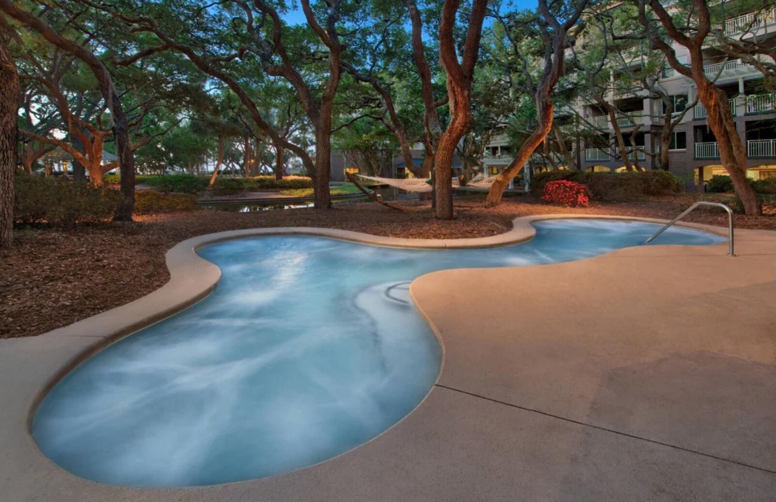 Une piscine au design incurvé, entourée d’arbres et bordée d’un bâtiment doté de balcons.