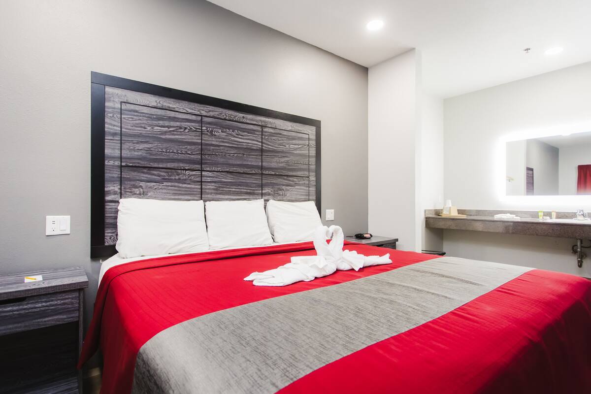 A hotel room with a large bed, a headboard, a bedside table, a nightstand, a mirror, and a bathroom counter.