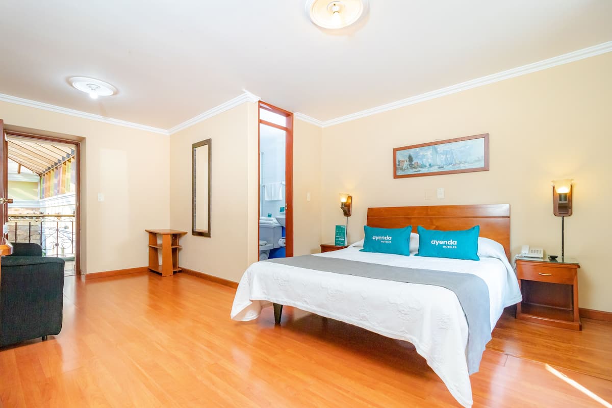 Ein Hotelzimmer mit einem großen Bett, Holzboden, Blick durch eine geöffnete Tür, einem Gemälde an der Wand und einem Nachttisch mit einer Lampe.