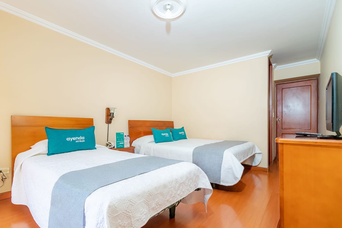 Ein Hotelzimmer mit zwei Betten, jedes mit einem türkisfarbenen Kopfkissen und einer weißen Decke. Es gibt ein hölzernes Kopfteil, einen Nachttisch mit Lampe sowie eine Holzkommode mit einem Fernseher.