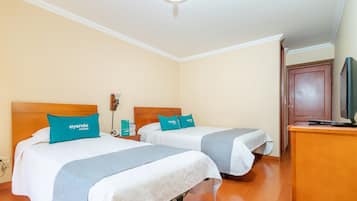 A hotel room with two beds, each with a teal pillow and a white blanket. There is a wooden headboard, a nightstand with a lamp, and a wooden dresser with a television.