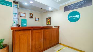 A hotel reception area with a wooden reception desk, a clock, framed certificates, and a sign for 'ayenda hoteles'.