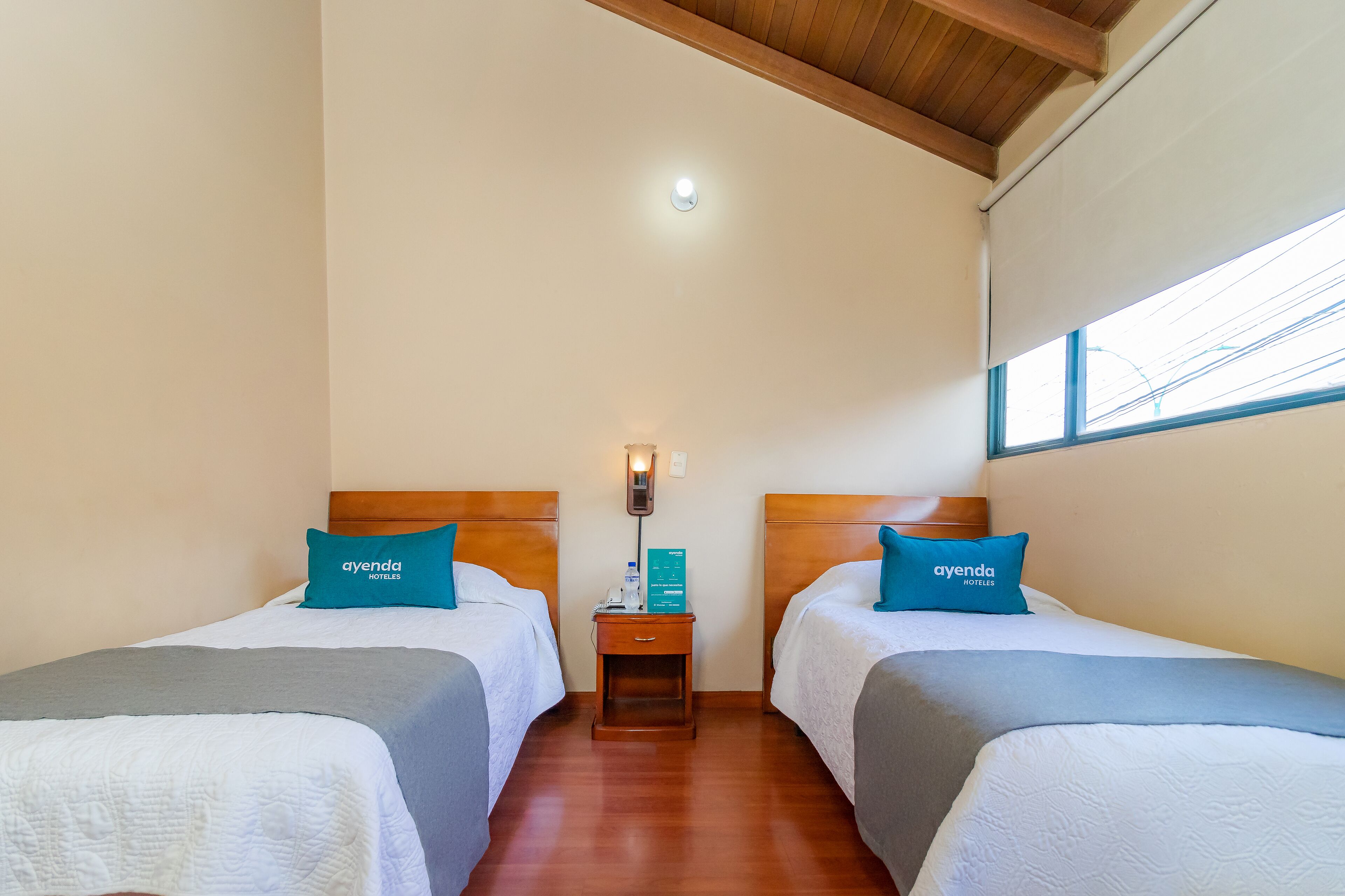 A hotel room with two beds, each with a blue pillow labeled 'ayenda hotels', a bedside table with a lamp, and a window with blinds.