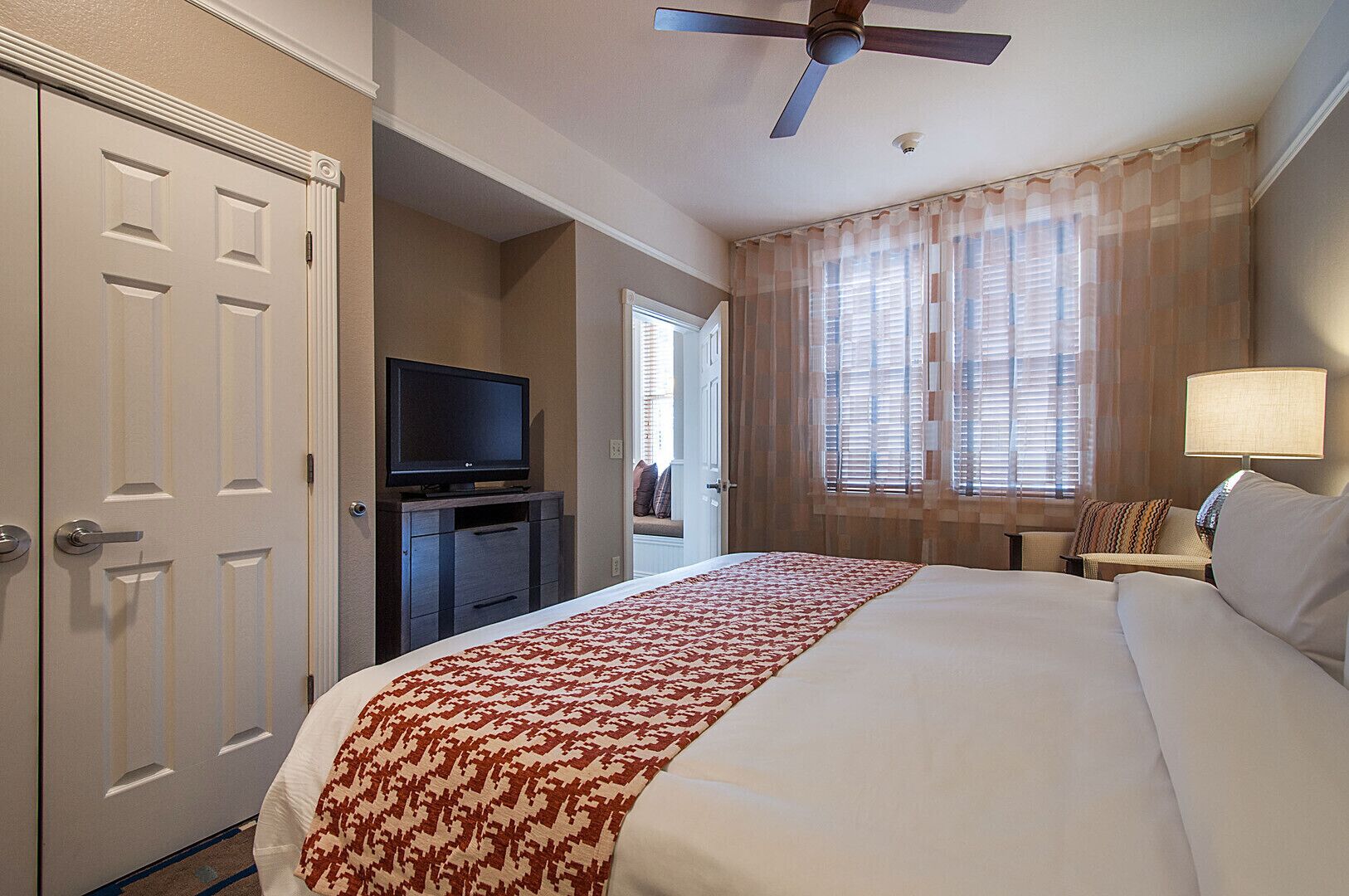 A hotel room with a large bed, a television on a stand, a ceiling fan, and a window with curtains.