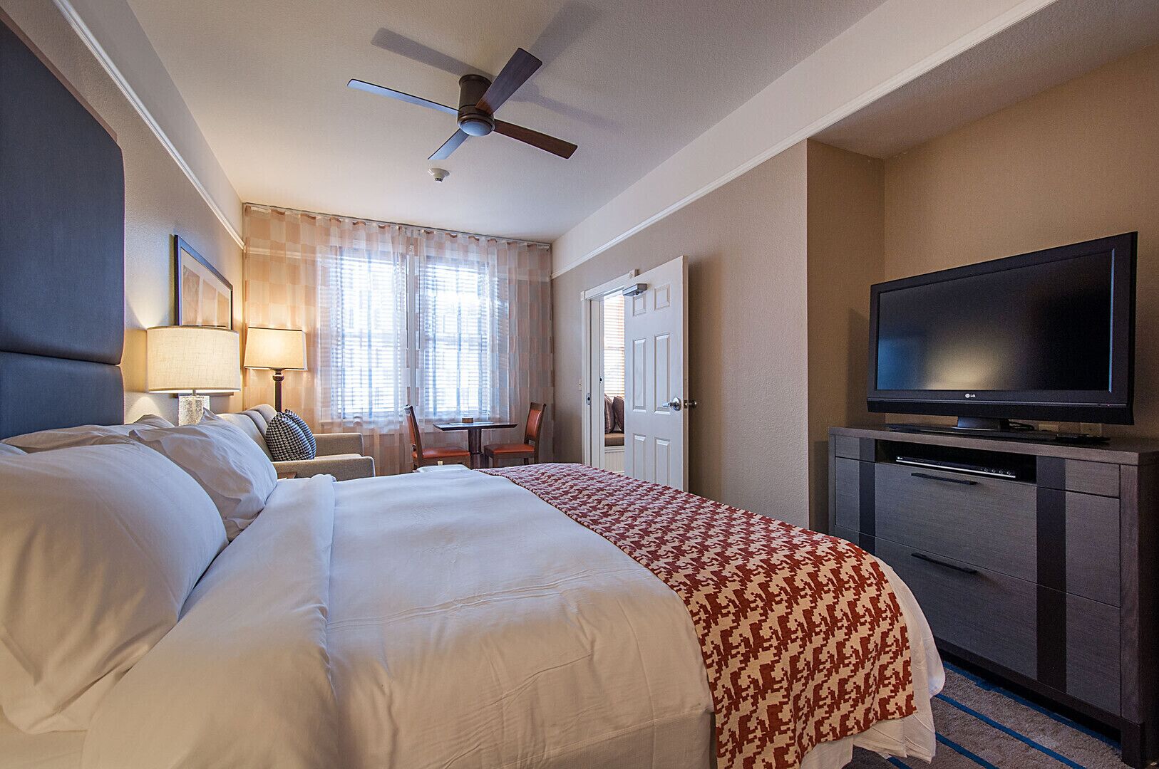 A hotel room with a bed, a desk, a chair, a television, and a ceiling fan.