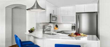 Eine moderne Küche mit weißen Schränken, einem Edelstahl-Kühlschrank, einer Mikrowelle und einem Spülbecken. Auf der Arbeitsplatte stehen zwei blaue Barhocker und eine Pflanze.