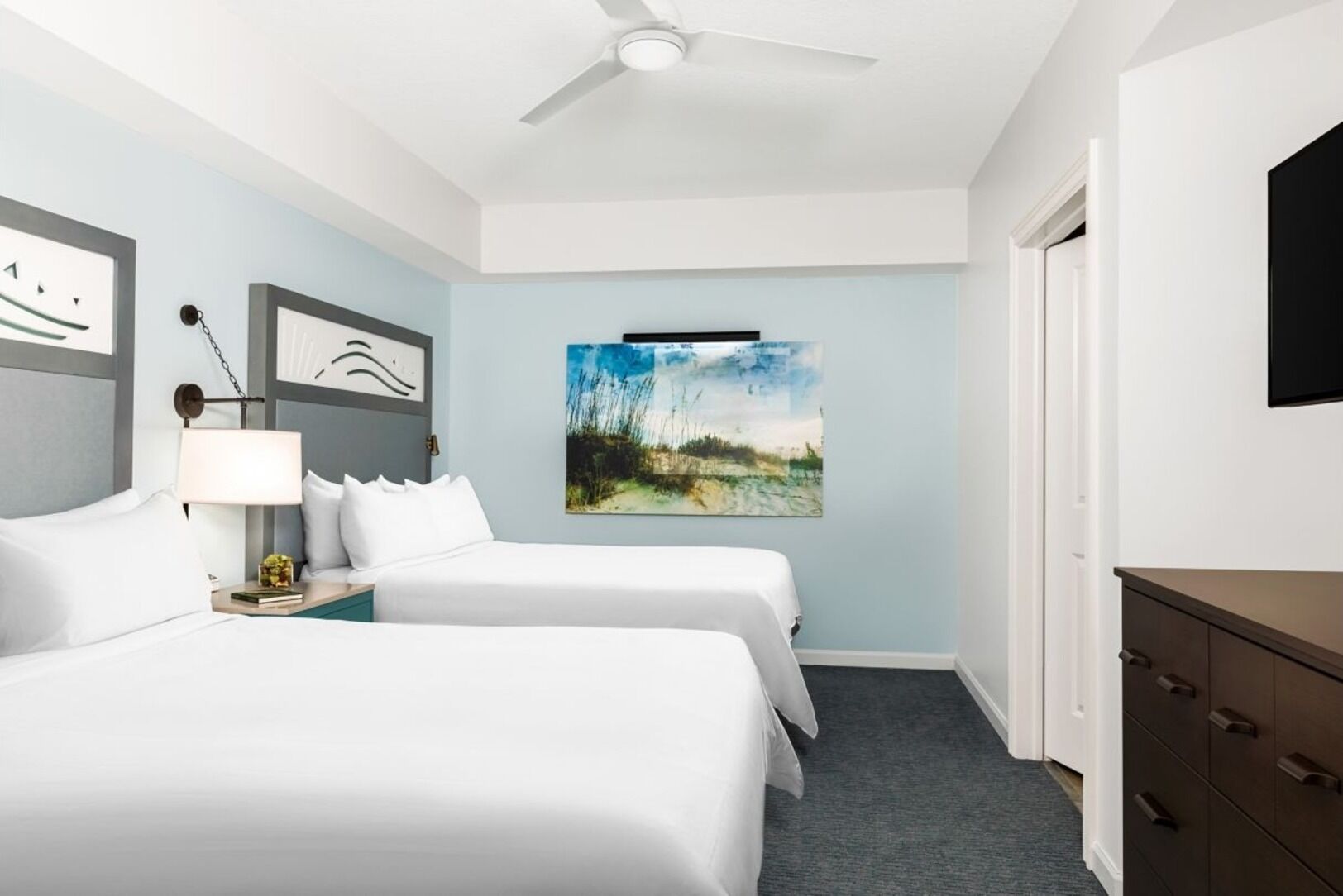 Una camera d'albergo con due letti, una ventola a soffitto, un quadro e un comò.