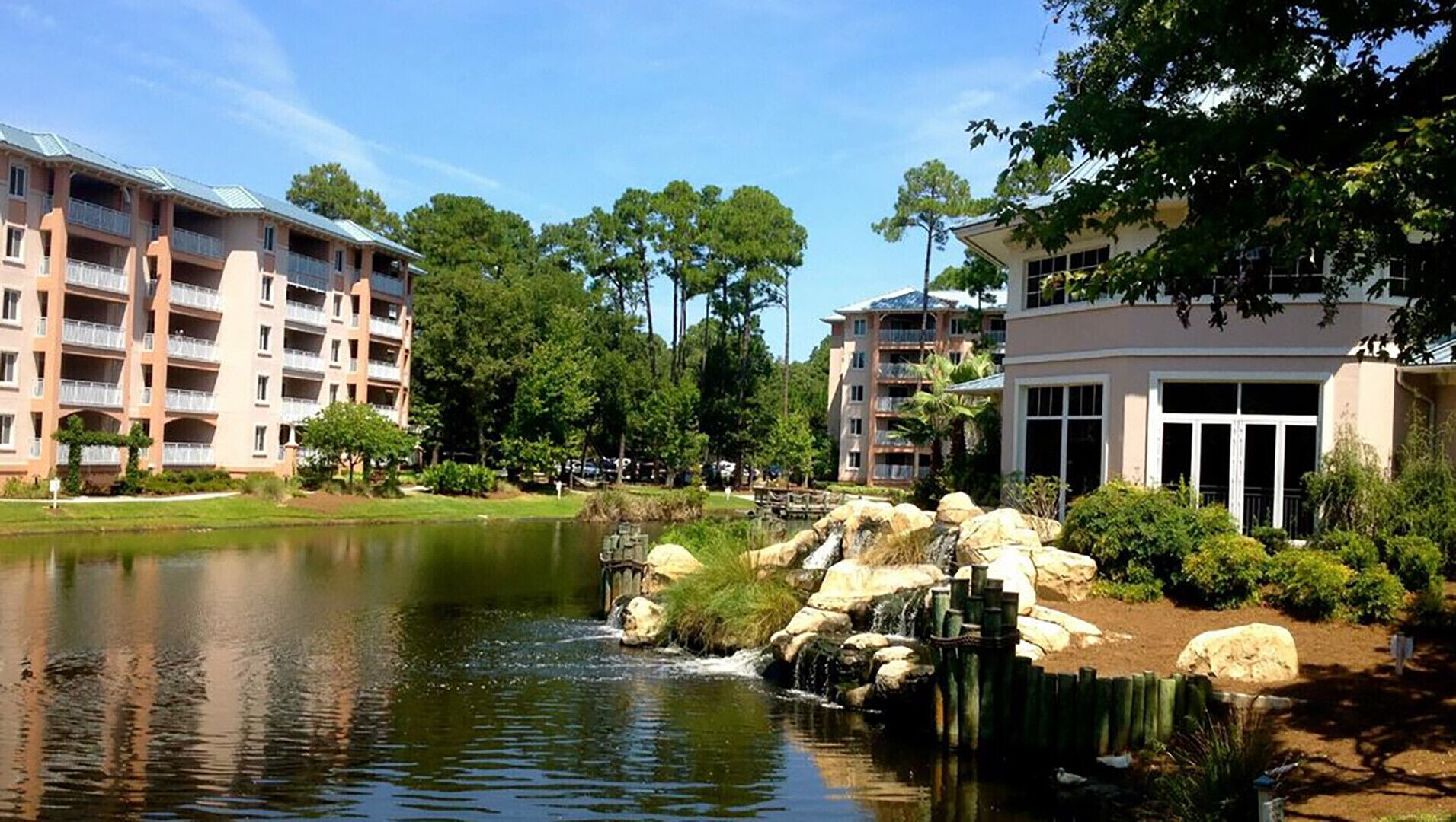 A lakeside view with a small waterfall, surrounded by multi-story buildings with balconies and a well-maintained garden area.