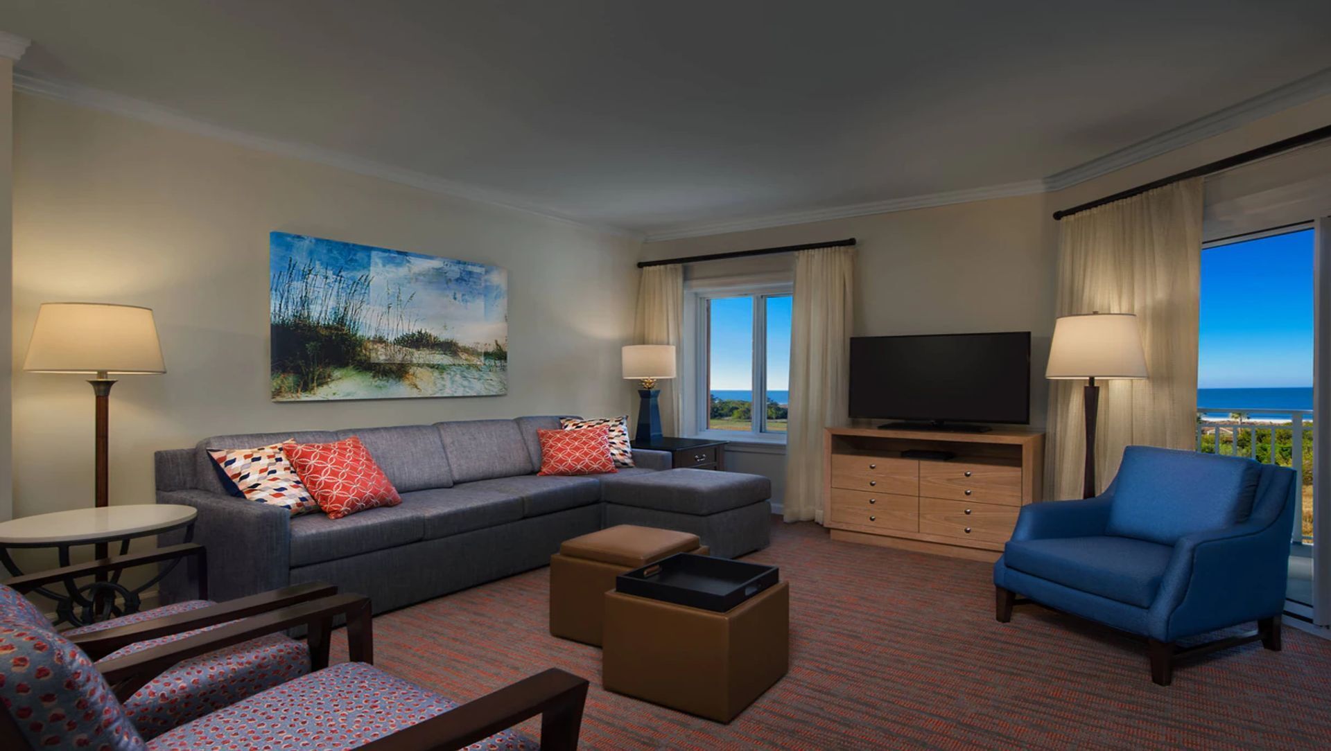 Ein Hotelzimmer mit grauem Sofa, blauem Sessel, Fernseher, Gemälde und Meerblick.