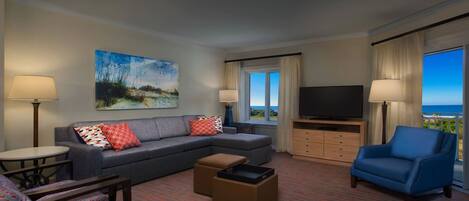 Ein Hotelzimmer mit grauem Sofa, blauem Sessel, Fernseher, Gemälde und Meerblick.