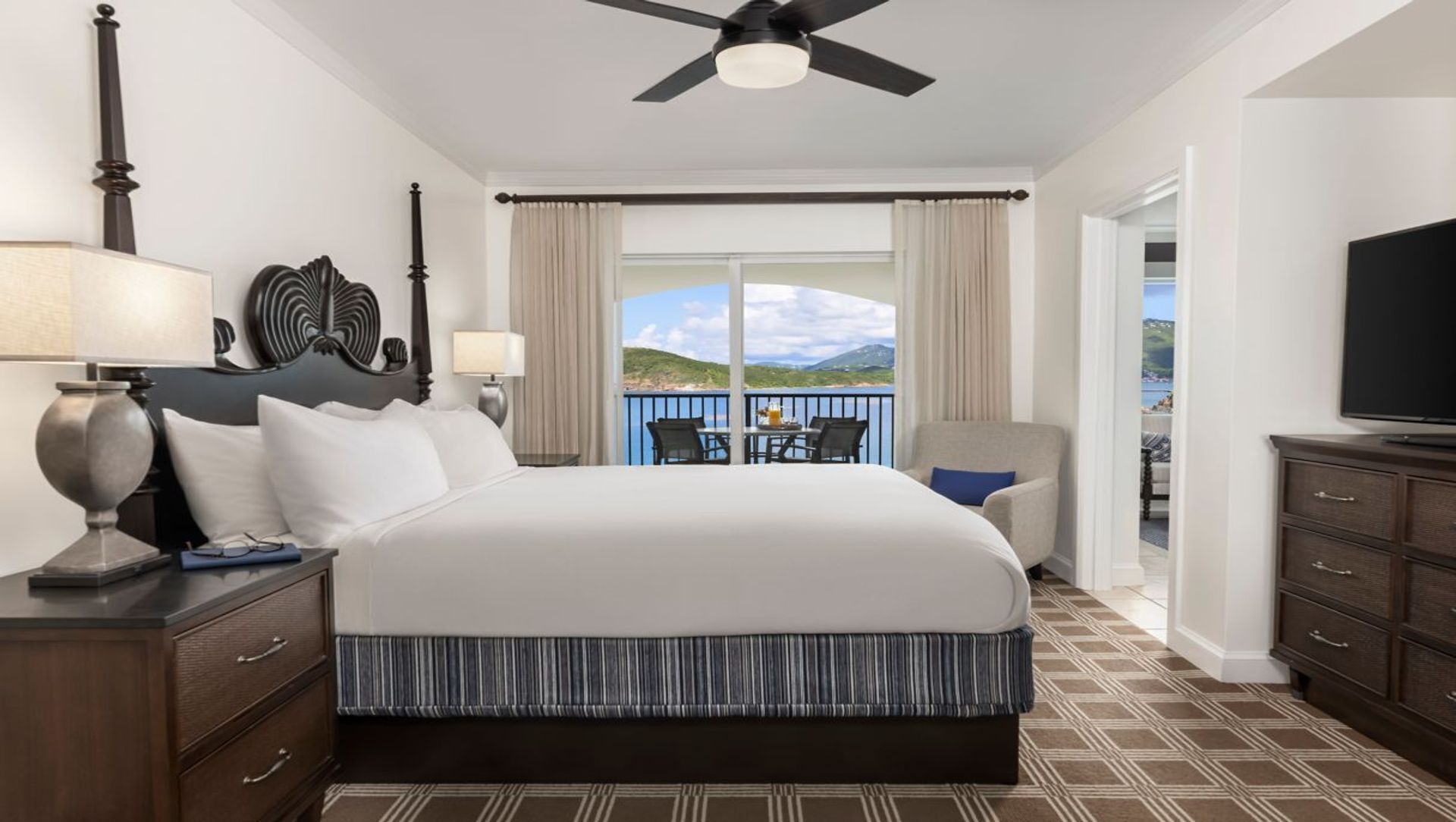 Habitación de hotel con una cama grande, ventilador de techo, televisión, balcón con vistas al paisaje y zona de comedor con sillas y mesa.