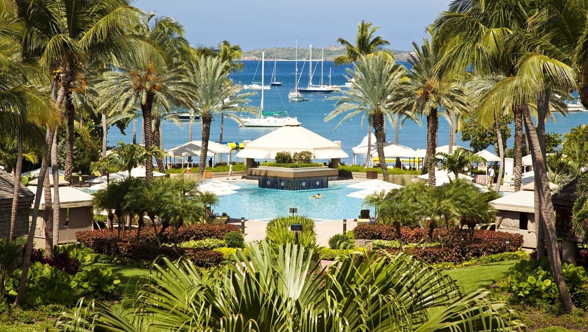 Un resort con piscina centrale, circondato da palme e vista sul mare con barche.