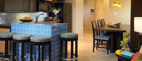 A modern kitchen with a blue tiled island, bar stools, a dining table with chairs, and a refrigerator.